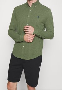 Mężczyzna w dopasowanej oliwkowej koszuli z guzikami i czarnych szortach do kolan, z rękami złożonymi na biodrach.