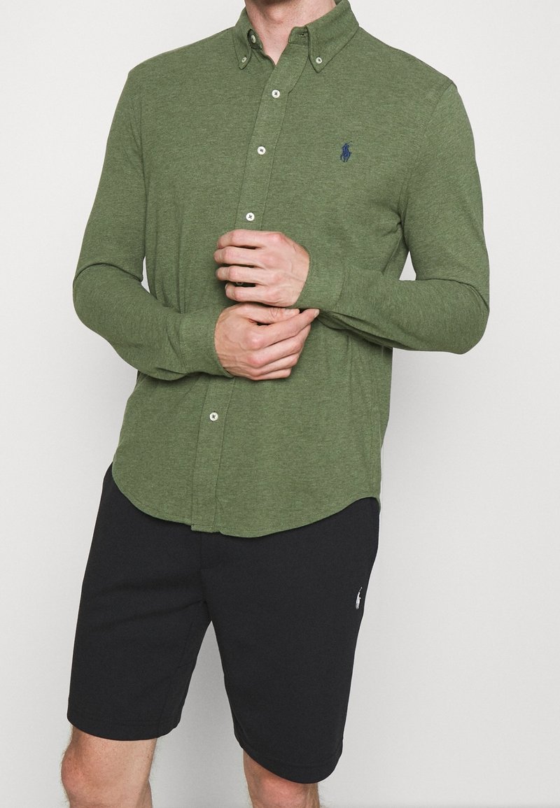 Mężczyzna w dopasowanej oliwkowej koszuli z guzikami i czarnych szortach do kolan, z rękami złożonymi na biodrach.