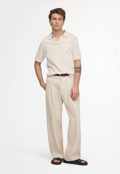 Homme aux cheveux bruns courts portant un polo beige, un pantalon ample beige, une ceinture noire, des sandales noires et un tatouage sur le bras, debout avec une main dans la poche.