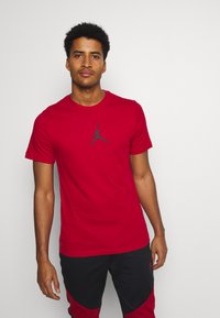 Rød bomulds t-shirt med korte ærmer, der har et sort Jumpman-logo centreret på brystet. Glat tekstur, rund halsdesign.