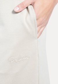 Lysebeige bukser i stof med sidelomme og broderet "Smiledex"-logo. Glat tekstur med afslappet pasform.