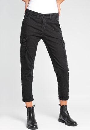 Nainen, jolla on mustat lyhyet cargohousut, joissa on napilliset sivutaskut, ja mustat nilkkurit, seisoo käsivarret lanteilla.