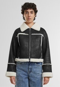 Veste noire courte avec un col et des poignets en fausse fourrure blanche, dotée d'une fermeture éclair et de coutures contrastées à l'avant.