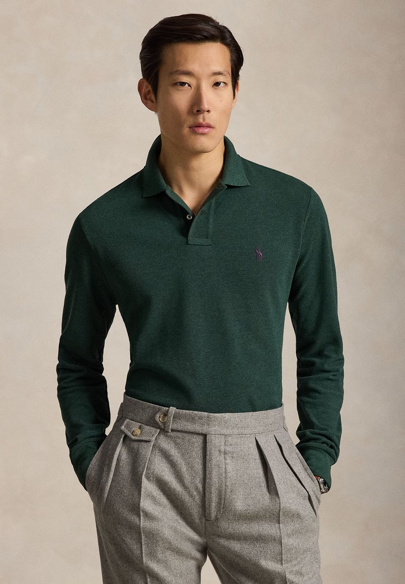 Man draagt een donker groen poloshirt met lange mouwen, ingestopt in hoog getailleerde grijze geplooide broek, handen in de zakken.