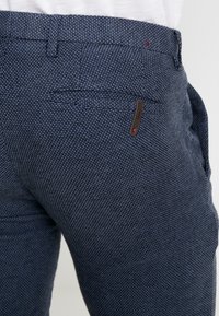 Navy blue trousers made from textured fabric, featuring a sleek fit, pocket detailing, and a small branded accent on the side pocket.