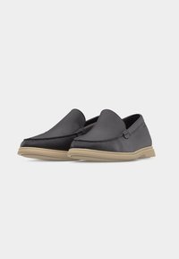 Paire de mocassins en cuir noir à enfiler avec semelles en caoutchouc beige, présentant un dessus cousu de style mocassin.
