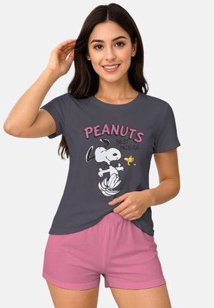 Junge Frau lächelt, trägt ein dunkelgraues Peanuts-T-Shirt mit Snoopy und Woodstock sowie eine rosa Shorts, vor einfachem hellem Hintergrund.