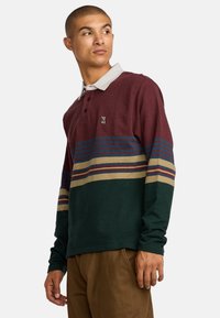 Camisa polo de manga larga en burdeos y verde, con patrones a rayas en azul, dorado y rojo. Incluye un cuello gris y un sutil detalle del logotipo.
