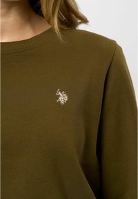 Temno olivno zelena dukserica z rebrastim okroglim izrezom. Ima majhen bel vezen logotip na levem prsnem delu. Mehka, teksturirana tkanina.