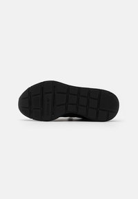 Semelle de chaussure noire avec un design en caoutchouc texturé, présentant des motifs de grip rectangulaires et circulaires, ainsi qu'un marquage en relief.