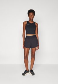 Top deportivo negro corto con un pequeño logo de acento, combinado con pantalones cortos negros que tienen una cintura elástica y bolsillos laterales. Zapatillas de correr negras.