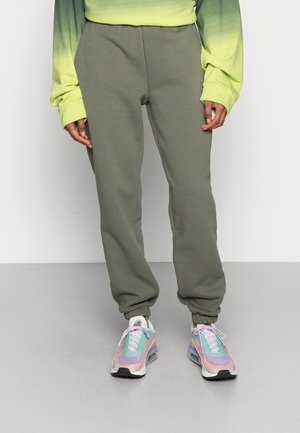 Osoba nosící olivově zelené teplákové kalhoty s elastickými manžetami, žlutozelený gradientní top s dlouhými rukávy a pastelové vícebarevné tenisky.