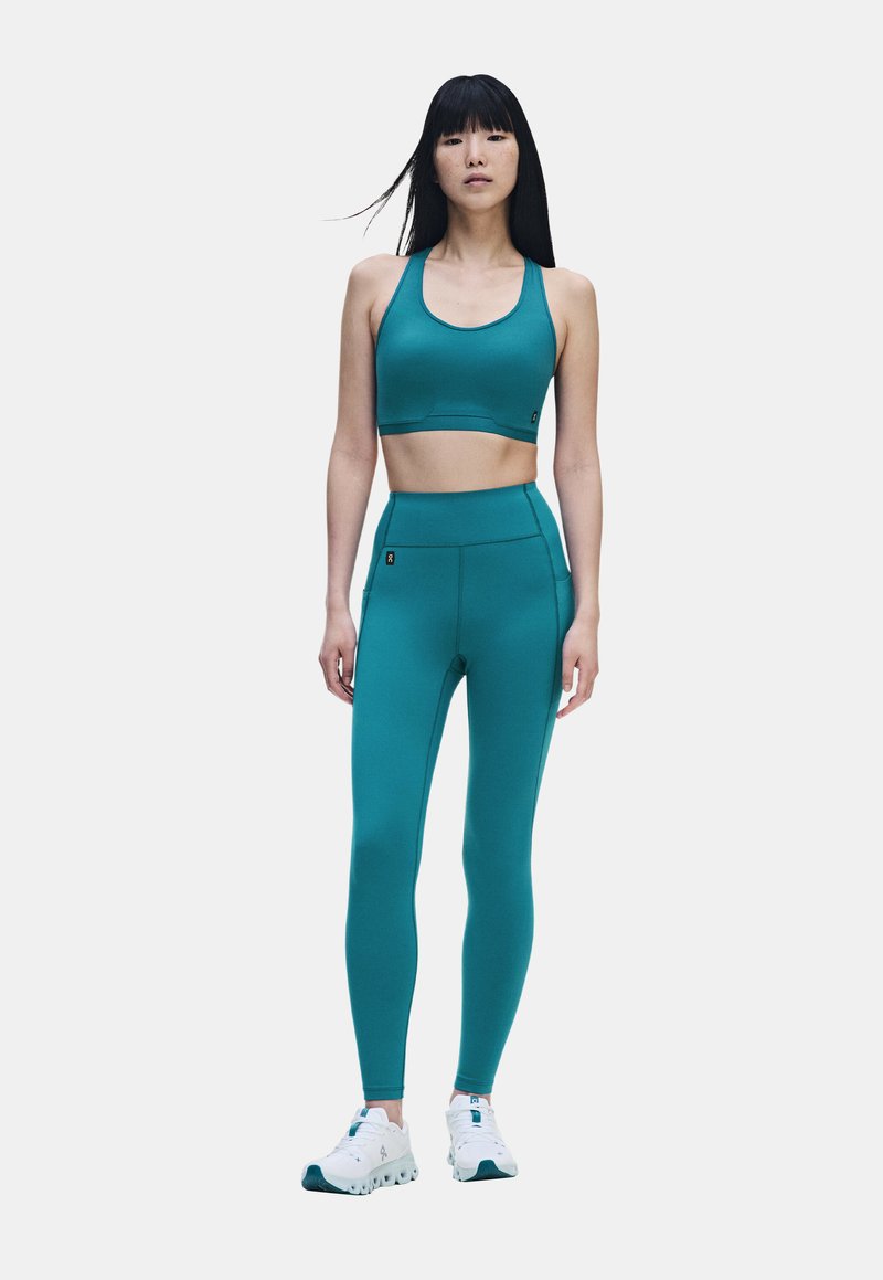 Femme aux longs cheveux noirs portant un soutien-gorge de sport bleu sarcelle et des leggings avec des chaussures de sport blanches, debout face à l'avant.