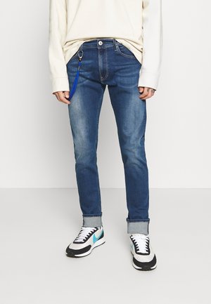 Person iført opsmøgede blå jeans, hvid langærmet skjorte, hvide og sorte sneakers med turkis swoosh og en blå nøglering fastgjort til jeansene.