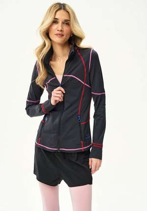 Kobieta o długich blond włosach, ubrana w czarną kurtkę na zamek z różowym i czerwonym przeszyciem, czarne krótkie spodenki i jasnoróżowe legginsy, stojąca na białym tle.