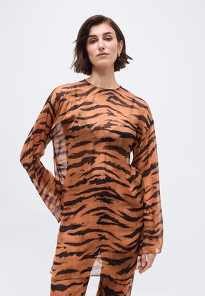 Femeie cu păr scurt și castaniu poartă un top transparent cu dungi în stil tigru și mâneci lungi, stând pe un fundal simplu, deschis la culoare.