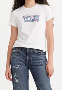 Bílá polokošile s krátkým rukávem s potiskem květinového loga Levi's na modrém pozadí, spárovaná s roztrhanými tmavomodrými džíny.