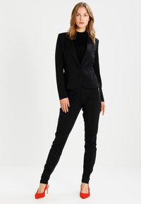 Svart skräddarsydd kavaj med en elegant design, enkelknappsstängning, tillsammans med svarta byxor och röda högklackade skor. Strukturerad tygfinish.