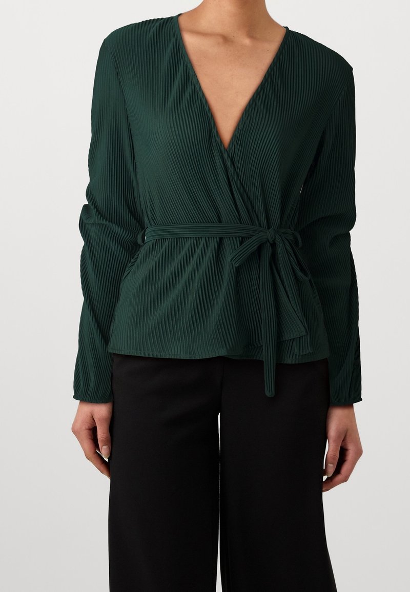 Donna in una blusa drappeggiata plissettata verde scuro con scollatura a V e cintura annodata, abbinata a pantaloni larghi neri, visibile dalle spalle ai fianchi.