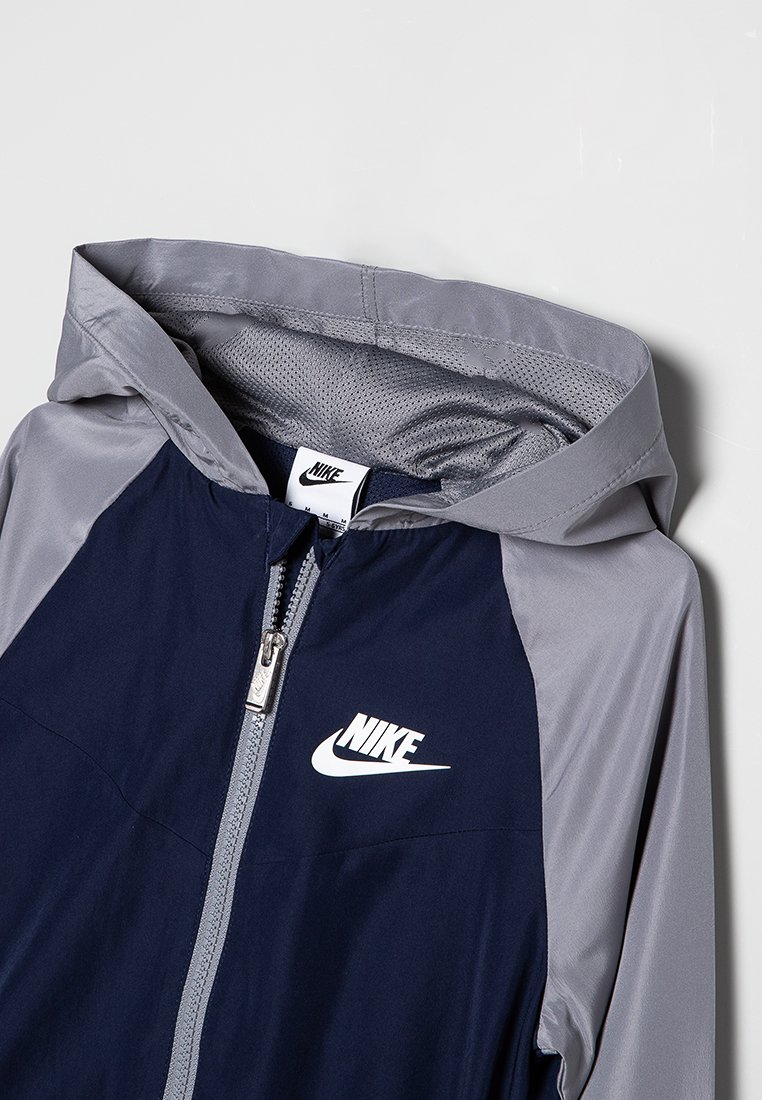 Giacca con zip navy e grigia con cappuccio, realizzata in tessuto leggero. Presenta un prominente logo Nike bianco sul petto.