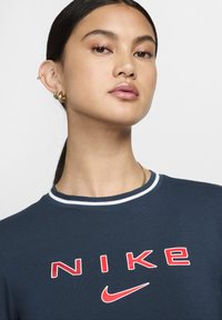 Tricou bleumarin cu guler ribbit alb și roșu; prezintă logo-ul "NIKE" în roșu îndrăzneț și un mic design al swoosh-ului roșu.