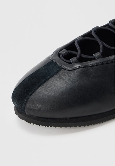 Μαύρο δερμάτινο παπούτσι με σουέντ πάνελ, στρογγυλεμένη μύτη και περίπλοκο σχέδιο κορδονιών στο πάνω μέρος. Διαθέτει υφασμένη λαστιχένια σόλα.