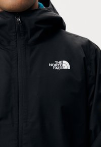 Veste imperméable noire avec un col montant, arborant le logo The North Face en blanc sur le côté gauche de la poitrine et une texture lisse et mate.