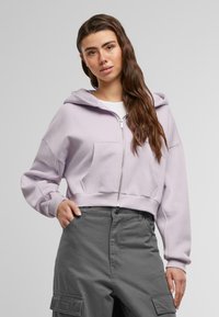 Lys lilla cropped hoodie med lynlås foran, to sidelommer og hætte med snor, parret med grå cargo bukser.