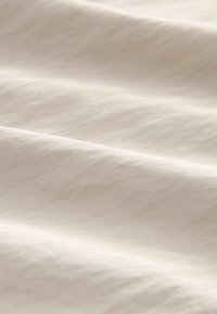 Tissu beige clair avec une texture lisse, affichant des plis doux et ondulés ainsi qu'un léger brillant, créant une apparence élégante et minimaliste.