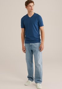 Kék pamut v-nyakú póló világoskék farmernadrággal párosítva. Az anyag sima, laza szabású. A tornacipő fehér, minimális részletezéssel.