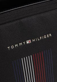 Tessuto nero texturizzato con il logo di Tommy Hilfiger in lettere bianche e un emblema rettangolare sopra strisce verticali rosse, bianche e blu.