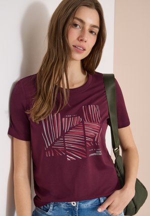Frau mit langen braunen Haaren, die ein lila T-Shirt mit Grafik und blaue Jeans trägt und eine grüne Umhängetasche dabei hat.