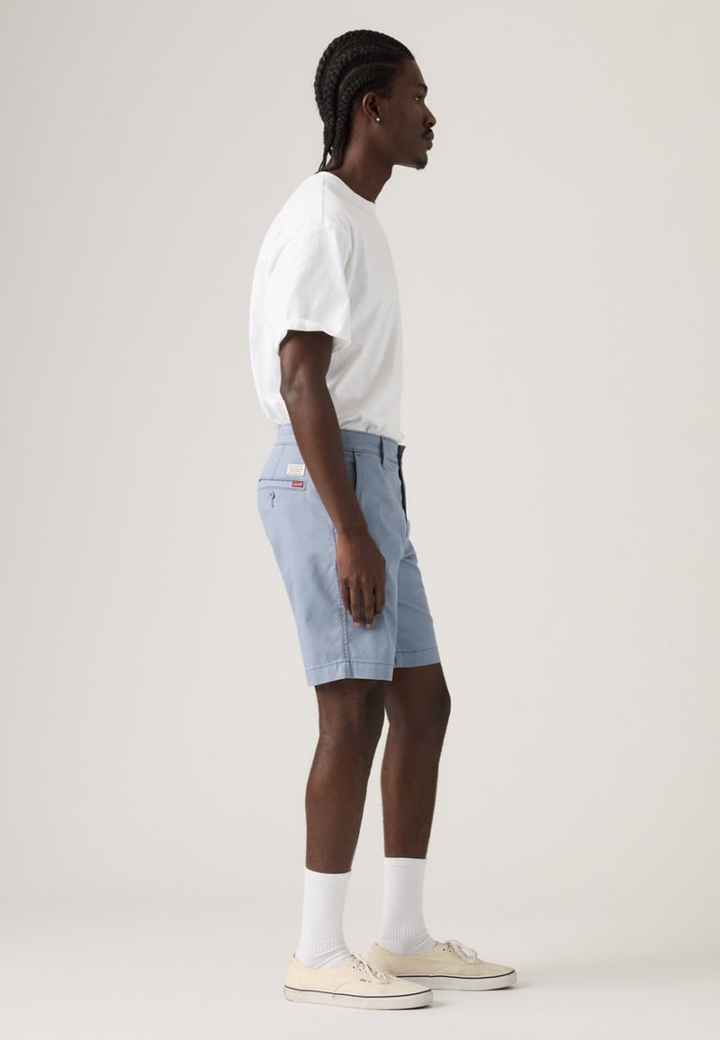 Homme aux cheveux tressés debout de profil, portant un t-shirt blanc, un short bleu clair avec un petit logo, des chaussettes blanches et des baskets beige.