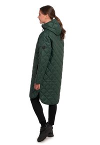Abrigo largo acolchado verde con capucha, que presenta un patrón de diamantes, cortes laterales y detalles negros. Lleva puesto sobre pantalones negros y calzado negro.