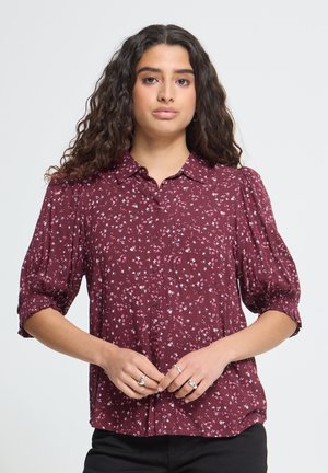 Femme aux longs cheveux bouclés portant un chemisier bordeaux à boutons, à petit motif fleuri et manches mi-longues, debout les mains jointes.