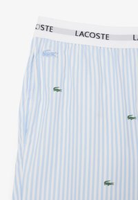 Lacoste blauwe en witte gestreepte shorts met een witte elastische tailleband met de merknaam. Groene krokodil logo geborduurd op de stof.