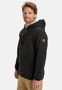 Felpa con zip di colore grigio scuro, con cappuccio foderato in pile, tasche frontali e dettagli in pelle marrone sulla zip e sulle maniche. Tessuto morbido e strutturato.