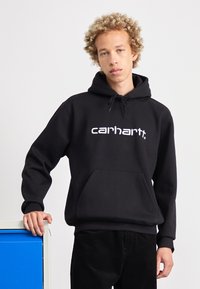 Svart hettegenser med lomme foran og hvit "Carhartt" logo. Glatt stofftekstur, avslappet passform, kombinert med mørke kordfløyelsbukser.