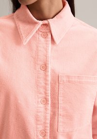 Roze corduroy overhemd met een klassieke kraag, borstzak en knoopsluiting aan de voorkant. De stof heeft een fijne verticale ribbelstructuur.