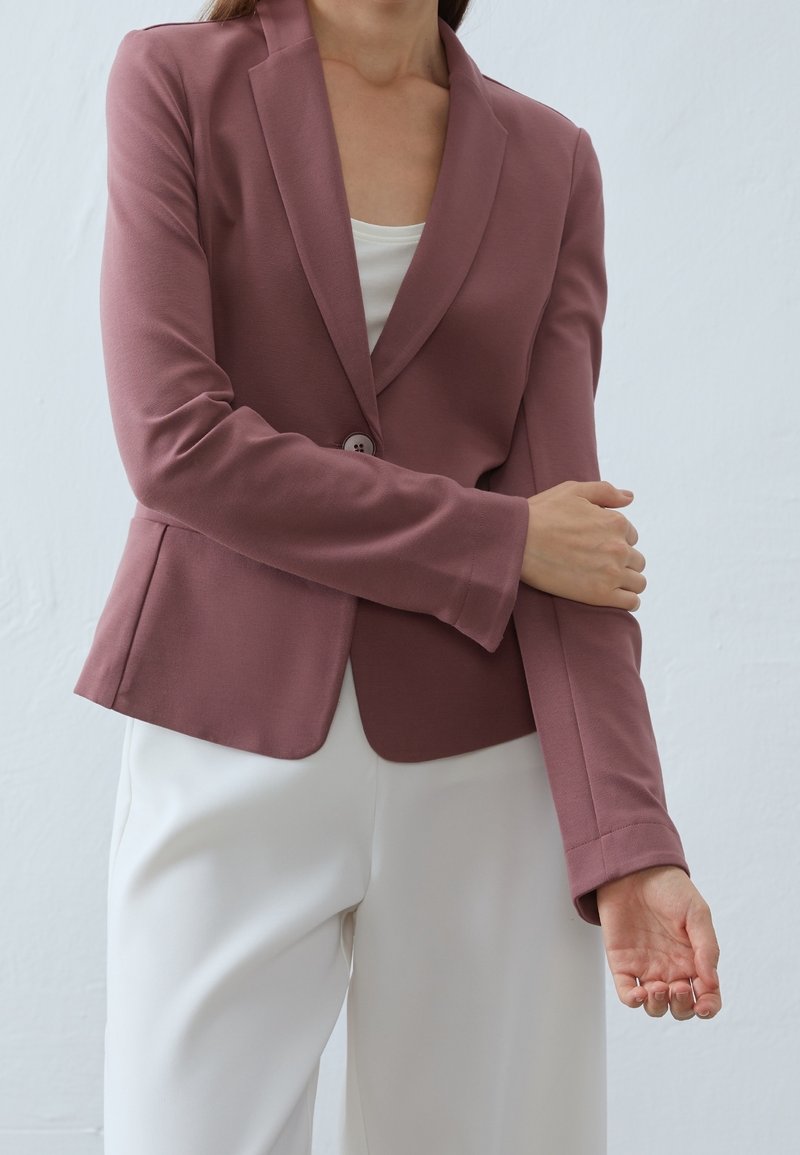Femme portant un blazer mauve par-dessus un top blanc et un pantalon blanc, ajustant la manche du blazer avec sa main gauche.