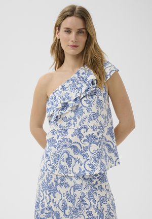 Nő fehér, egyrészes vállpántos felsőt és hozzáillő, kék virágos paisley mintás szoknyát visel, vállán fodros részlettel.