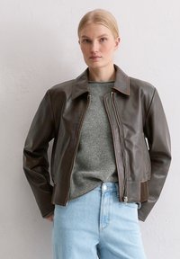 Bruine leren jas met een recht design, opvallende kraag, ritssluiting en contrasterende stiksels, gedragen over een grijze trui en blauwe spijkerbroek.