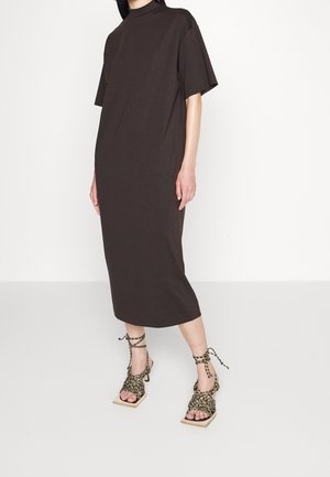 Persoon draagt een loszittende donkerbruine jurk tot op de knie met korte mouwen en een hoge halslijn, gecombineerd met patroonrijke veterhakken met open teen.