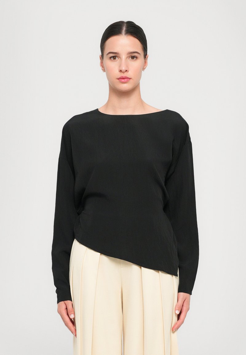 Black long-sleeve top with an asymmetrical hem, featuring a round neckline and smooth, lightweight fabric. Paired with beige pleated trousers.