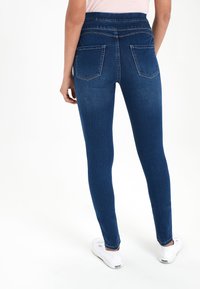 Jean skinny en denim bleu foncé avec une taille haute. Comprend deux poches arrière et une texture lisse. Associé à des baskets blanches.