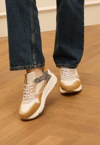 Sneakers met een beige bovenkant, beige en tan suède stukken, accenten met luipaardprint en dikke witte zolen. G gedragen met donkere denim.