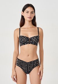 Donna che indossa un set di lingerie trasparente nera con pois bianchi, in piedi contro uno sfondo grigio chiaro uniforme.