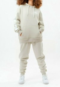 Personne portant un sweat à capuche beige oversize et un pantalon de jogging assorti avec des baskets blanches, mains dans la poche du sweat, debout devant un fond blanc.