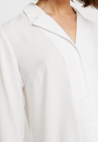 White button-up shirt with a classic collar, made of smooth fabric, featuring subtle texture and a relaxed fit with pearl-like buttons.