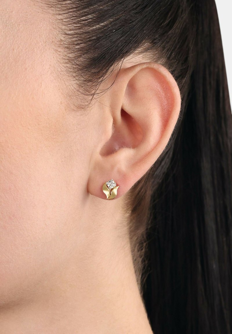Orecchio di donna con capelli lisci e scuri raccolti dietro, che indossa un piccolo orecchino a bottone d'oro a forma di fiore con una pietra centrale trasparente.